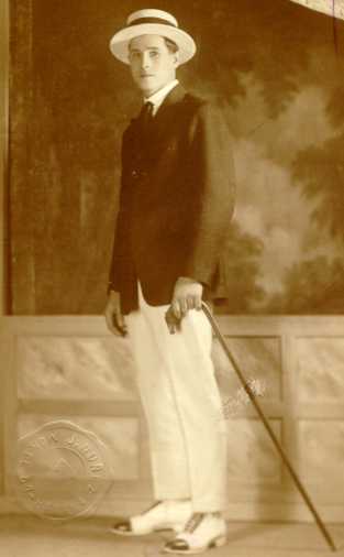 Foto que muestra a un joven indiano (20 años) con botines blancos, pantalón blanco, chaqueta oscura con corbata y sombrero de paja de la época, con bastón en la mano.